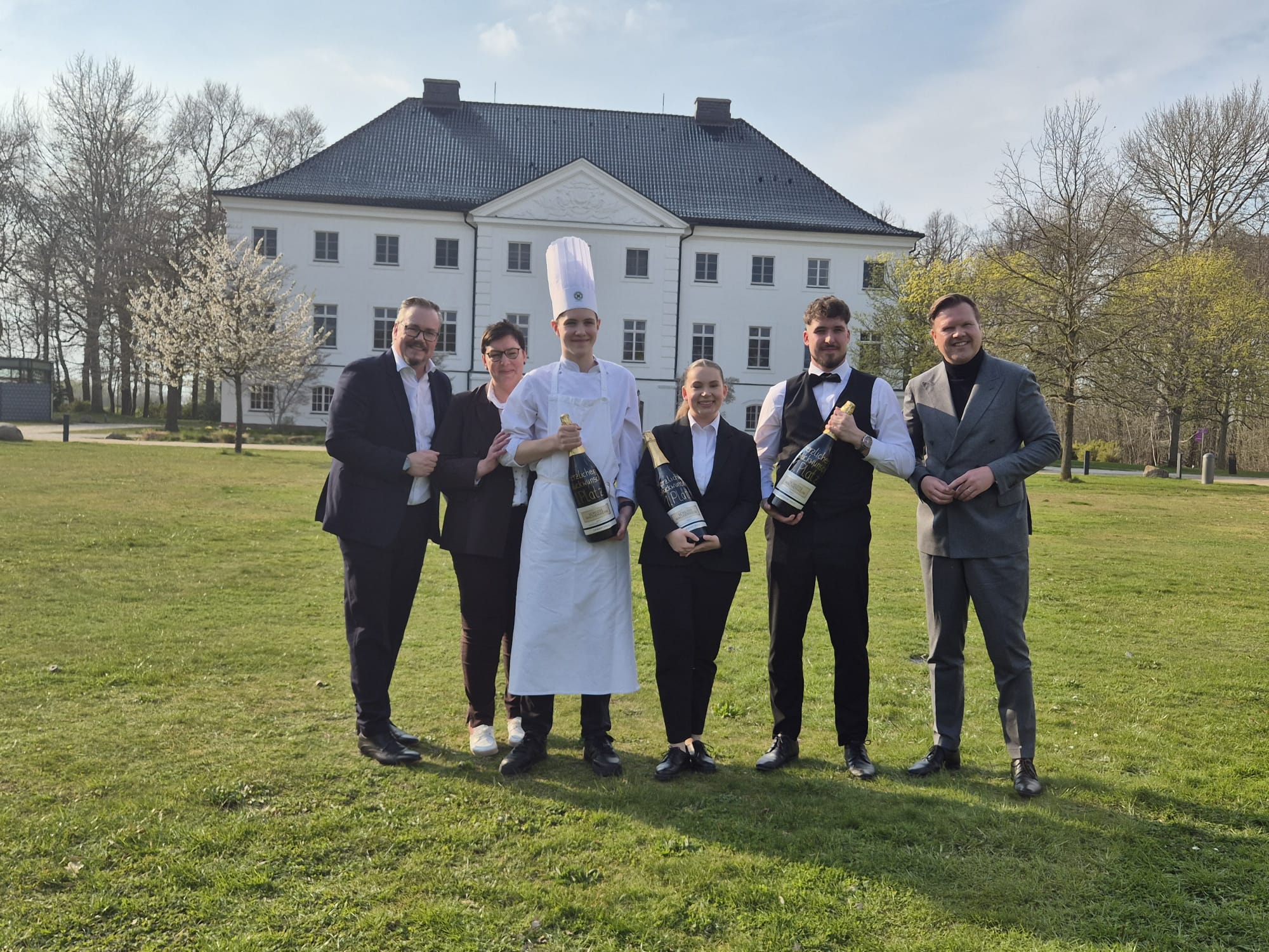 Die Gewinnerin und der Gewinner des Wettbewerbs halten Champagnerflaschen in den Händen. Neben Ihnen steht ein Teil des Veranstaltungsteams. Alle posieren auf einer grünen Rasenfläche mit dem Hotelgebäude im Hintergrund.