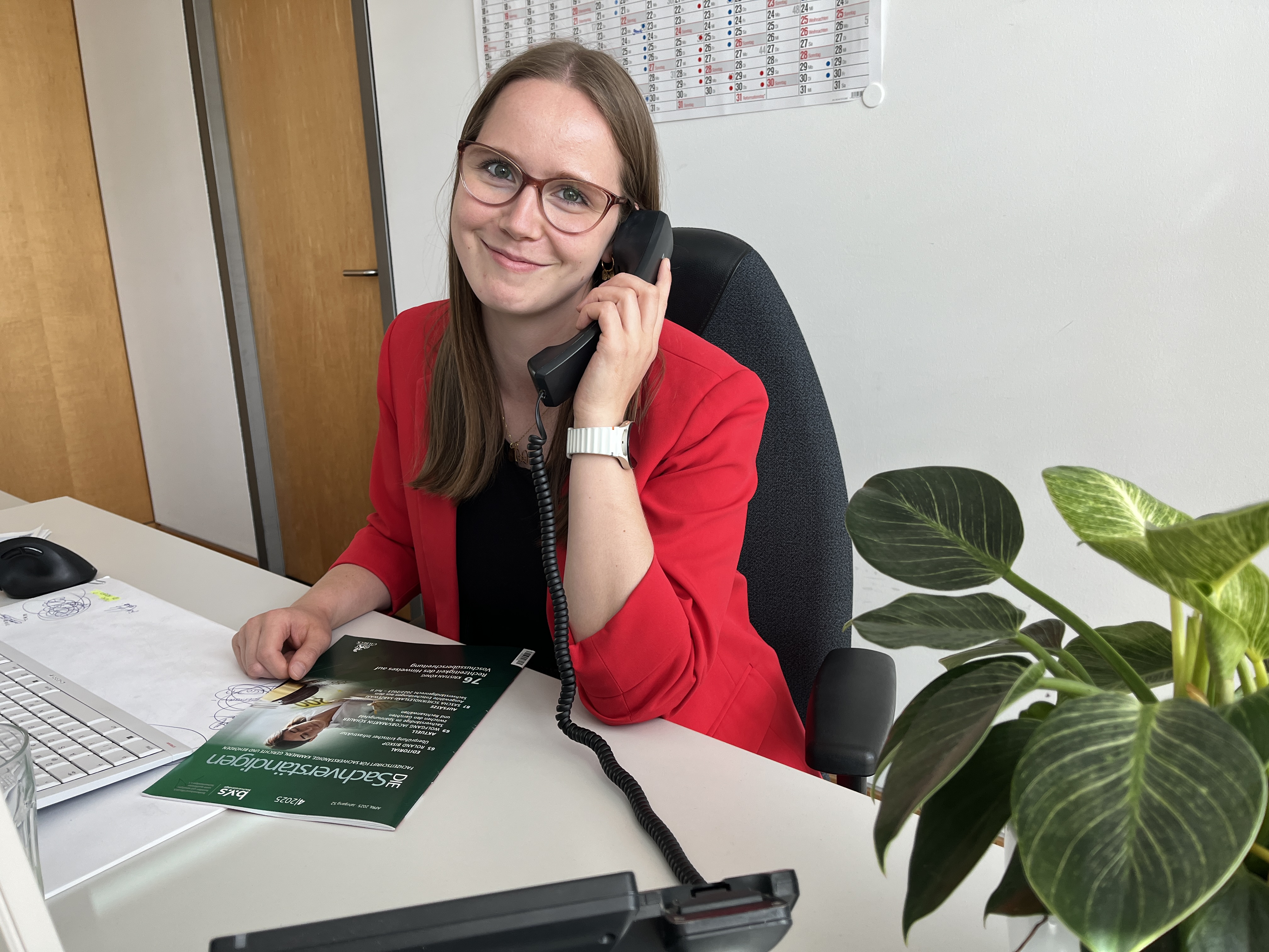 Annika Bünz sitzt an ihrem Schreibtisch, hält einen Telefonhörer an ihr Ohr und lächelt in die Kamera. 